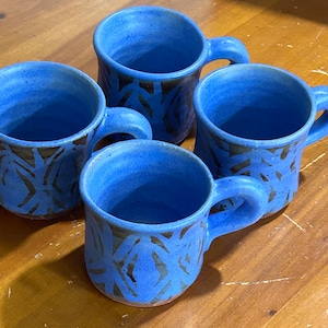 May include: Four handmade blue ceramic mugs with brown leaf-like patterns. Each mug has a rounded handle and a speckled finish. The mugs are arranged on a wooden surface, showcasing their unique design and craftsmanship.