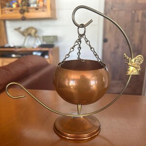 May include: A copper hanging planter with a round bowl and a decorative stand. The stand has a curved arm and a small windmill design.