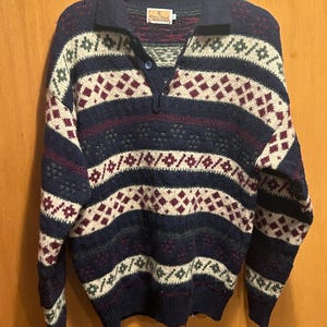 May include: A navy blue sweater with a collared neckline and a button closure. The sweater features a geometric pattern in cream, burgundy, and green. The sweater is hanging on a wooden hanger.