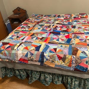May include: A colorful patchwork quilt with a variety of fabrics and patterns, including floral, geometric, and plaid designs. The quilt is laid out on a bed with a green floral bed skirt.