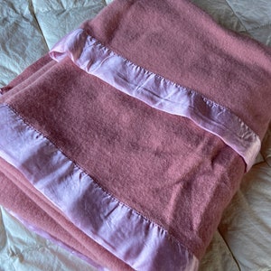 May include: A pink wool blanket with a light pink satin trim. The blanket is folded and lying on a white bedspread.