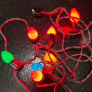 May include: A string of Christmas lights with red and green wiring. The lights are various colors, including red, orange, blue, and green. The lights are shaped like bulbs and are lit up. A black power cord is visible.