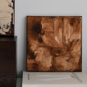 Könnte beinhalten: Quadratisches abstraktes Kunstwerk in Braun- und Schwarztönen. Die strukturierte Oberfläche zeigt Pinselstriche, eingefasst in einem dunklen Holzrahmen. Das Kunstwerk liegt auf einem Bücherstapel.