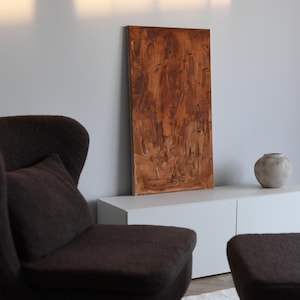 Könnte beinhalten: Ein braunes abstraktes Gemälde mit strukturierter Oberfläche lehnt an einem weißen Schrank. Im Vordergrund steht ein dunkelbrauner Sessel mit passendem Kissen. Eine kleine, runde, graue Vase steht auf dem Schrank.