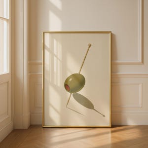 Könnte beinhalten: Ein gerahmter vertikaler Wandkunstdruck, der eine einzelne grüne Olive mit einem roten Pimentkern zeigt, die von einem dünnen hölzernen Zahnstocher aufgespießt ist. Das Bild ist auf hellem cremefarbenem Grund in einem dünnen Goldrahmen.