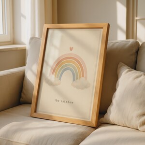 Op de afbeelding: Een ingelijste kunstprint met een pastelkleurige regenboog met wolken aan beide uiteinden en een klein hartje erboven. De tekst "the rainbow" is in het zwart onderaan in het midden gedrukt op de crèmekleurige achtergrond in een lichte houten lijst.