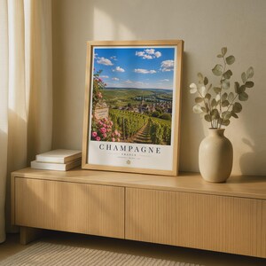 May include: A framed vertical wall art print featuring a scenic landscape of vineyards in Champagne, France, with pink flowers in the foreground, a blue sky with clouds, and the text "CHAMPAGNE FRANCE" printed in black at the bottom of the white border.