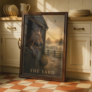 Op de afbeelding: Een ingelijste rechthoekige wandkunstprint met een bruin paard dat bij zonsopgang uit een houten stalbox kijkt. Een zadel hangt aan de staldeur naast een hooinet. De tekst THE YARD is in het wit onderaan de afbeelding gedrukt.