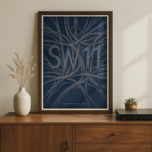 Puede incluir: Una lámina artística enmarcada con fondo azul oscuro que presenta intrincadas ilustraciones de líneas blancas de vías de tren que forman las letras SW11. El texto THE JUNCTION - SW11 está impreso en blanco en la parte inferior del diseño.