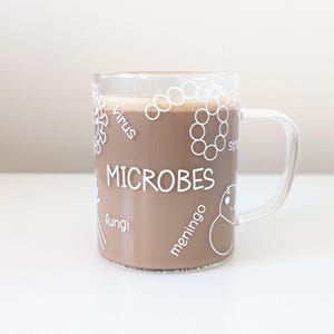 May include: A clear glass mug containing a brown beverage. The mug is decorated with a white outline of various microbes, including a virus, fungi, and bacteria, with the word "MICROBES" printed in white.