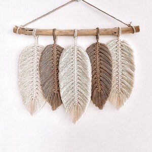 Op de afbeelding: Een wanddecoratie met vijf macramé veren in beige en bruine tinten, hangend aan een natuurlijke tak. De veren zijn met een beige koord aan elkaar gebonden en hangen aan een witte haak.