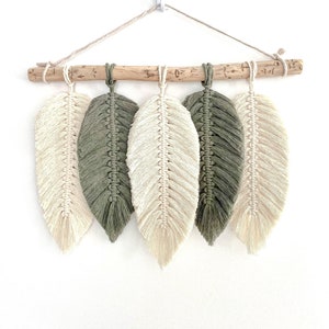 May include: Macrame feather wall hanging featuring five feathers in cream and olive green hues. The feathers are suspended from a natural driftwood branch with a neutral-colored cord for hanging. A decorative piece for a bohemian or nature-inspired aesthetic.