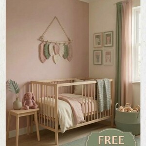 Puede incluir: Una cuna de madera clara con ropa de cama en color crema y rosa empolvado, una manta de punto verde salvia, un tapiz de macramé con plumas, una mesa auxiliar con un conejo de peluche y una cesta de almacenamiento verde salvia.