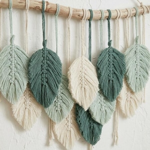 May include: A macrame wall hanging featuring multiple leaf-shaped fiber elements suspended from a natural wooden branch. The leaves are crafted in varying shades of cream, sage green, and dark forest green, arranged in a layered, overlapping pattern.
