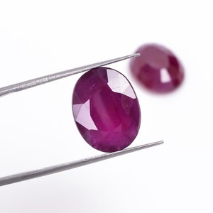 May include: A close-up of a deep red oval-shaped gemstone held by tweezers. The gemstone is faceted and has a high polish.