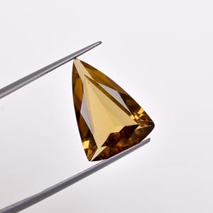 Peut inclure: Une pierre précieuse triangulaire facetée de couleur brun doré. La pierre précieuse est tenue par des pinces.