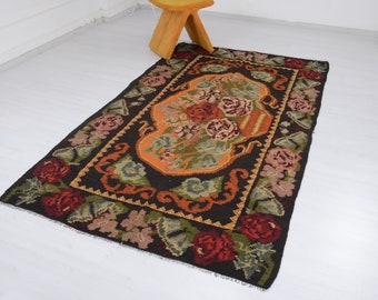 May include: A vintage Turkish rug with a floral design in shades of red, orange, green, and brown. The rug has a dark brown background and a central motif of a vase filled with flowers.