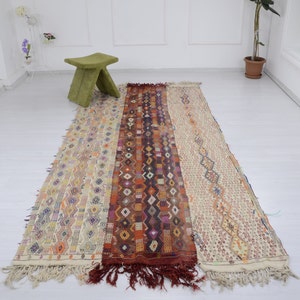 May include: A long, rectangular rug with a cream background and intricate geometric patterns in shades of orange, red, blue, and purple. The rug has a fringe on both ends.