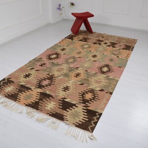 May include: A traditional Turkish kilim rug with a geometric pattern in shades of brown, beige, pink, and green. The rug has a fringe border and is woven with a variety of textures.