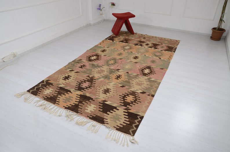 May include: A traditional Turkish kilim rug with a geometric pattern in shades of brown, beige, pink, and green. The rug has a fringe border and is woven with a variety of textures.
