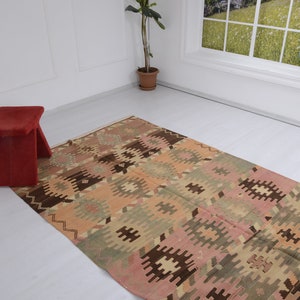 May include: A vintage Turkish kilim rug with a geometric pattern in shades of brown, pink, and green. The rug is laid out on a white floor with a red stool in the foreground.