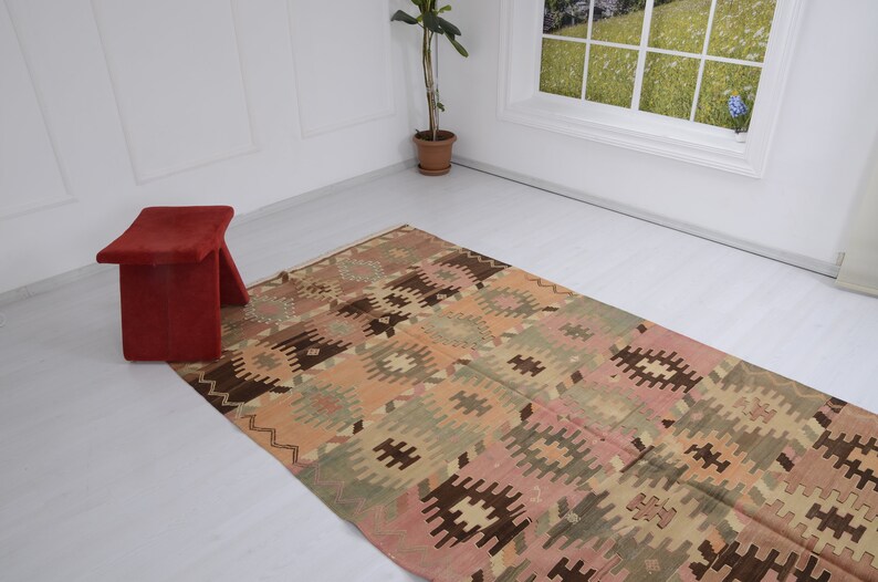 May include: A vintage Turkish kilim rug with a geometric pattern in shades of brown, pink, and green. The rug is laid out on a white floor with a red stool in the foreground.