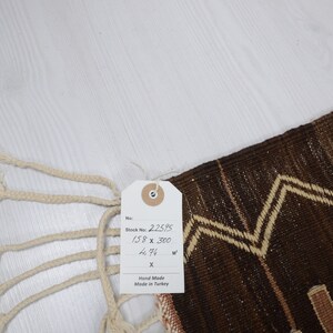 May include: A brown and cream handwoven rug with a geometric pattern. The rug has a tag attached that reads "Stock No. 2.25.95 158 x 300 4.94 sq m Hand Made Made in Turkey".