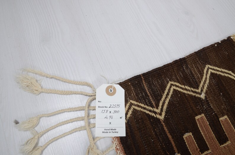 May include: A brown and cream handwoven rug with a geometric pattern. The rug has a tag attached that reads "Stock No. 2.25.95 158 x 300 4.94 sq m Hand Made Made in Turkey".