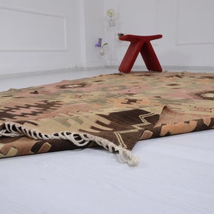 May include: A folded, hand-woven rug with a geometric pattern in shades of brown, beige, and pink. The rug has a fringe border and is lying on a white floor.
