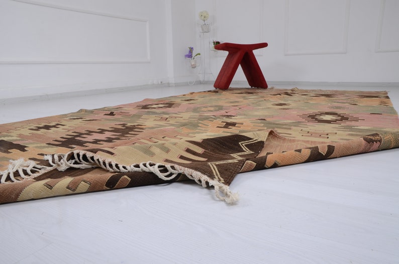 May include: A folded, hand-woven rug with a geometric pattern in shades of brown, beige, and pink. The rug has a fringe border and is lying on a white floor.