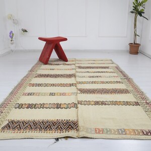 May include: A cream-colored rug with a geometric pattern in shades of brown, red, and green. The rug has a fringe border and is laid out on a white floor.