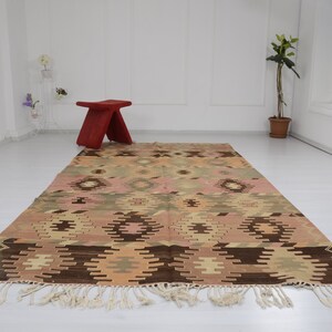 May include: A vintage Turkish kilim rug with a geometric pattern in shades of brown, beige, pink, and green. The rug has a fringe border and is laid out on a white floor.