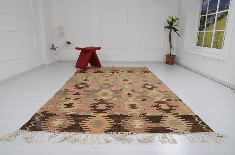 May include: A vintage Turkish kilim rug with a geometric pattern in shades of brown, beige, pink, and green. The rug has a fringe border and is laid out on a white floor.