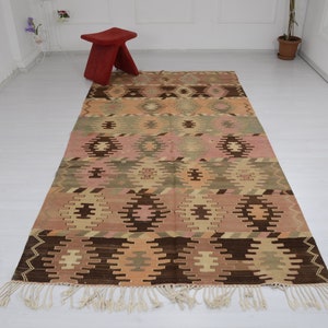 May include: A vintage Turkish kilim rug with a geometric pattern in shades of brown, pink, and beige. The rug has a fringe border and is woven with a traditional kilim technique.