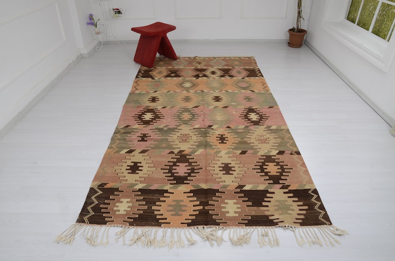 May include: A vintage Turkish kilim rug with a geometric pattern in shades of brown, pink, and beige. The rug has a fringe border and is woven with a traditional kilim technique.