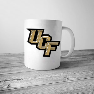 Pu&ograve; includere: Una tazza da caff&egrave; in ceramica bianca con un logo UCF nero e oro.