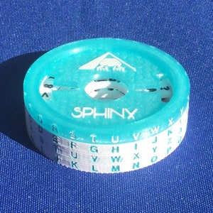 Könnte beinhalten: Eine türkisfarbene Chiffrierscheibe aus Kunststoff mit dem Wort "SPHINX" auf der Oberseite. Die Scheibe hat mehrere Schichten mit Buchstaben des Alphabets, die auf jeder Schicht gedruckt sind.