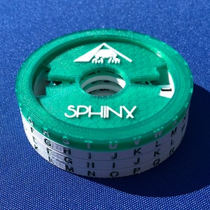 May include: A green and white plastic cipher disk with the word "SPHINX" printed on the top. The disk has multiple layers with letters of the alphabet printed on each layer.