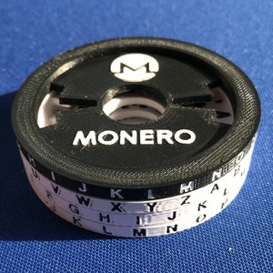 Könnte beinhalten: Eine in Schwarzweiß 3D-gedruckte Monero-Münze mit einem weißen Buchstaben 'M' auf der Oberseite und dem Wort 'MONERO' auf der Seite. Die Münze hat eine Reihe konzentrischer Ringe mit darauf gedruckten Buchstaben.