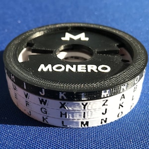 May include: A black and white 3D printed Monero cipher wheel with the letters of the alphabet printed on each ring. The top ring has the letter "M" printed in the centre.