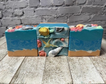 May include: Three bars of handmade soap with a beach theme. Each bar features a different ocean-inspired design with a seashell, starfish, and turtle. The soap is a light blue colour with a white swirl and a peach-coloured base.