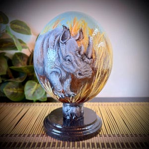May include: An oval-shaped decorative object featuring a hand-painted gray rhinoceros standing in tall yellow grass against a light blue sky. The piece is mounted on a circular black pedestal base, creating a smooth and polished display item.