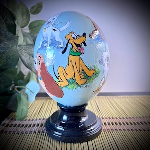 May include: A hand-painted ceramic egg with a blue background and images of Disney dogs, including Pluto, Bolt, and Lady. The egg is on a black stand.