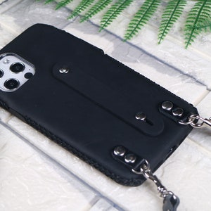 May include: A black phone case with a built-in phone grip and a black and silver phone strap. The case has a textured surface and a clear camera lens cover.