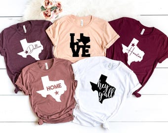May include: Five t-shirts with different Texas-themed designs. The shirts are in various colors, including maroon, beige, white, and a darker shade of pink. The designs feature the outline of the state of Texas with different text, including "Dallas", "Houston", "Love", "Home", and "Hey Y'all".