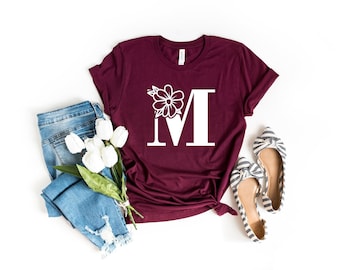 May include: A maroon t-shirt with a white floral design and the letter "M" in a large font.