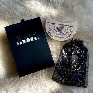May include: A black box with a gold inscription that reads "You are loved" and a crescent moon design. A wooden crescent moon with a bird and floral design and the text "You are loved" is also included. A black mesh bag with gold stars and moons is included.