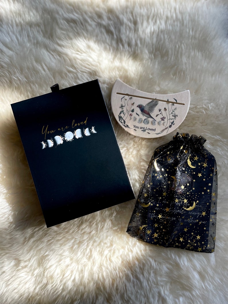 May include: A black box with a gold inscription that reads "You are loved" and a crescent moon design. A wooden crescent moon with a bird and floral design and the text "You are loved" is also included. A black mesh bag with gold stars and moons is included.