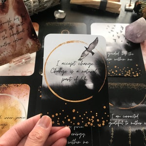 May include: A black and white affirmation card with gold accents. The card features a black bird flying against a white background with a gold circle around the text "I accept change. Change is a natural part of life."
