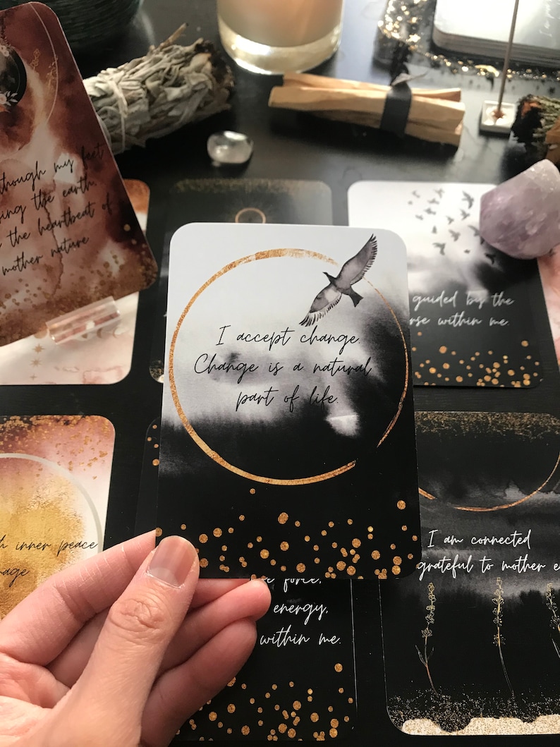 May include: A black and white affirmation card with gold accents. The card features a black bird flying against a white background with a gold circle around the text "I accept change. Change is a natural part of life."